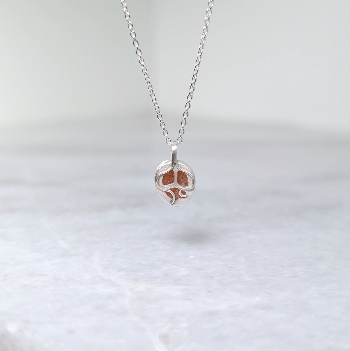 The back side of a silver pendant with an orange speckled stone visible through windows framed by silver vine motifs