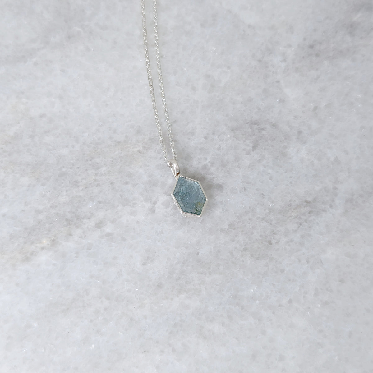 Silver necklace with a speckled light blue hexagonal stone set in a silver bezel, on a white marble surface