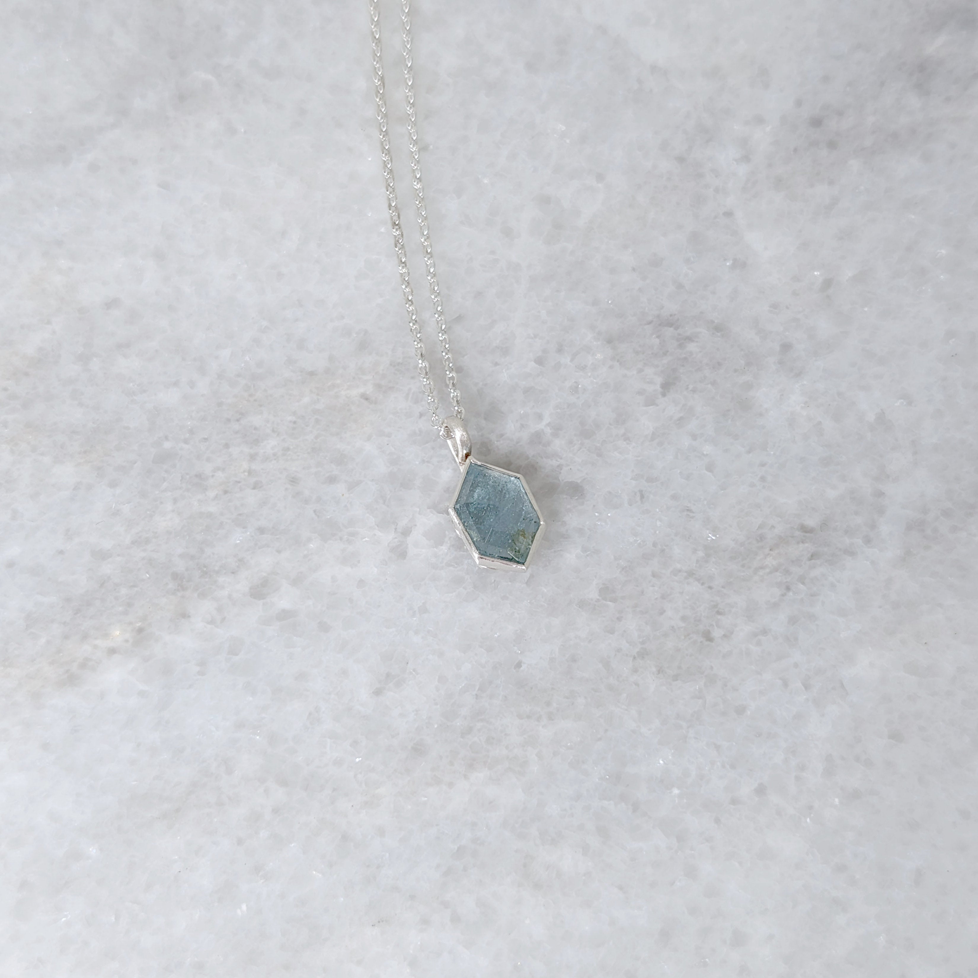 Silver necklace with a speckled light blue hexagonal stone set in a silver bezel, on a white marble surface