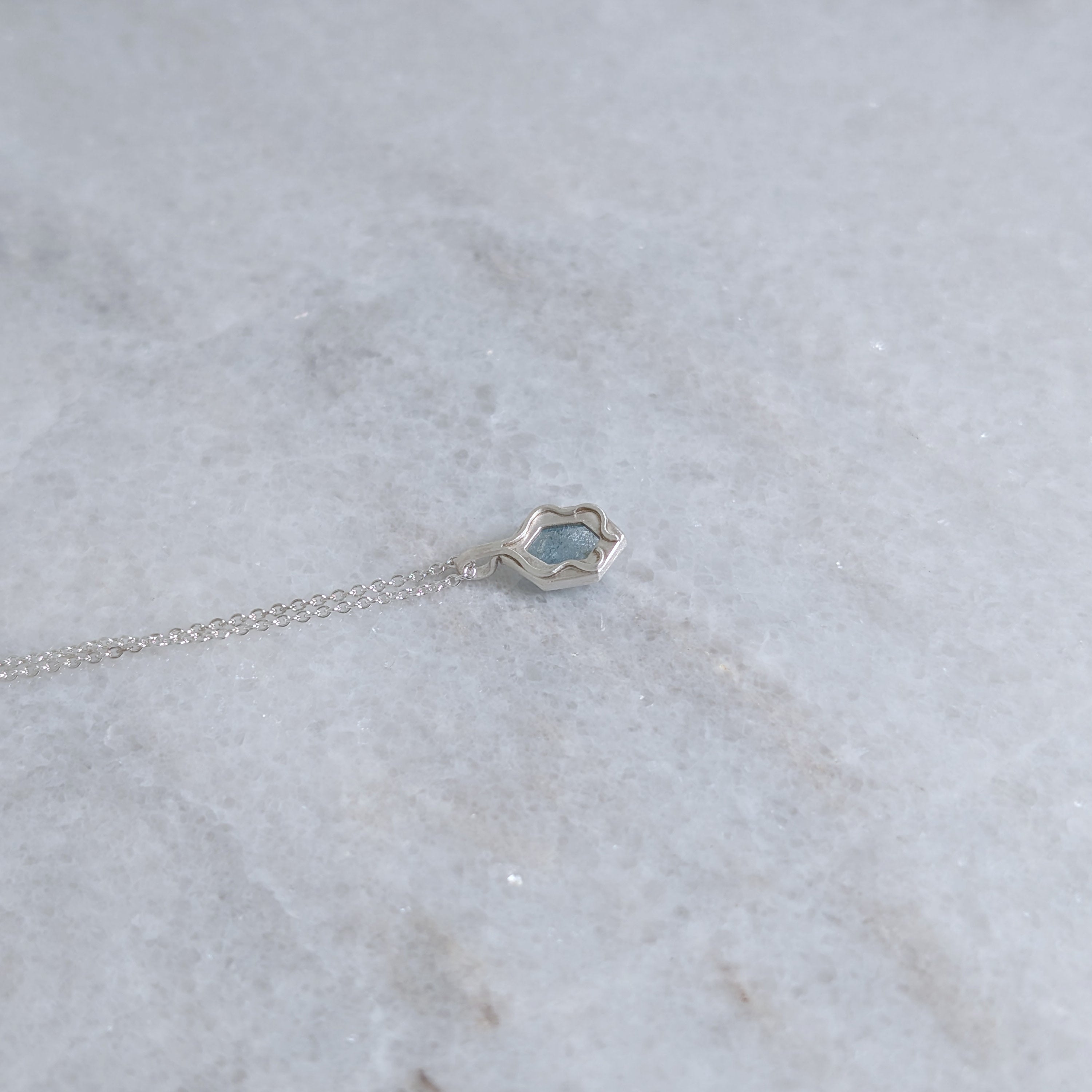 Back view of silver bezel with a windowed view of the light blue speckled stone with two vines framing it on a white marble sruface
