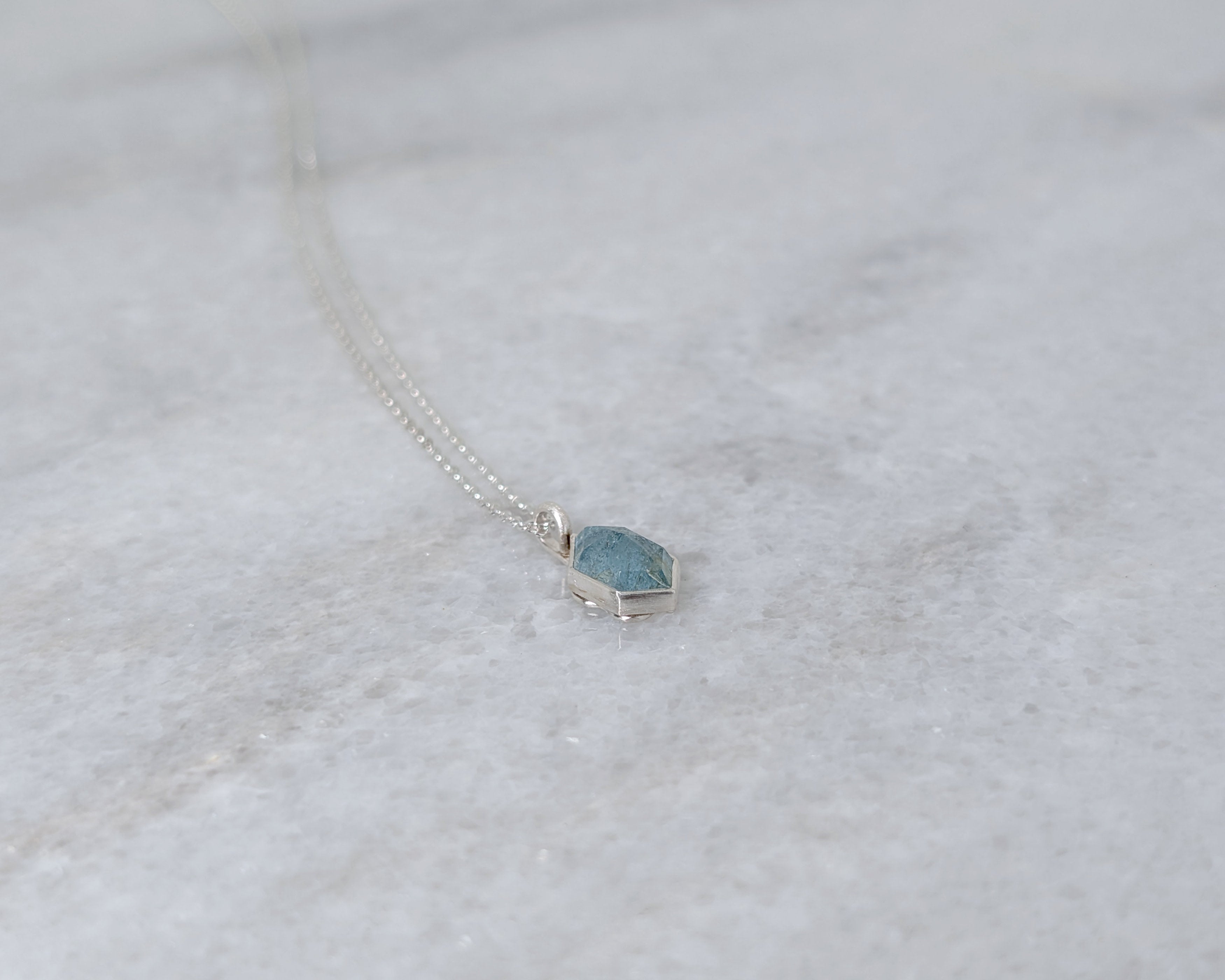 Angled view of a silver necklace with a speckled light blue hexagonal stone set in a silver bezel, on a white marble surface