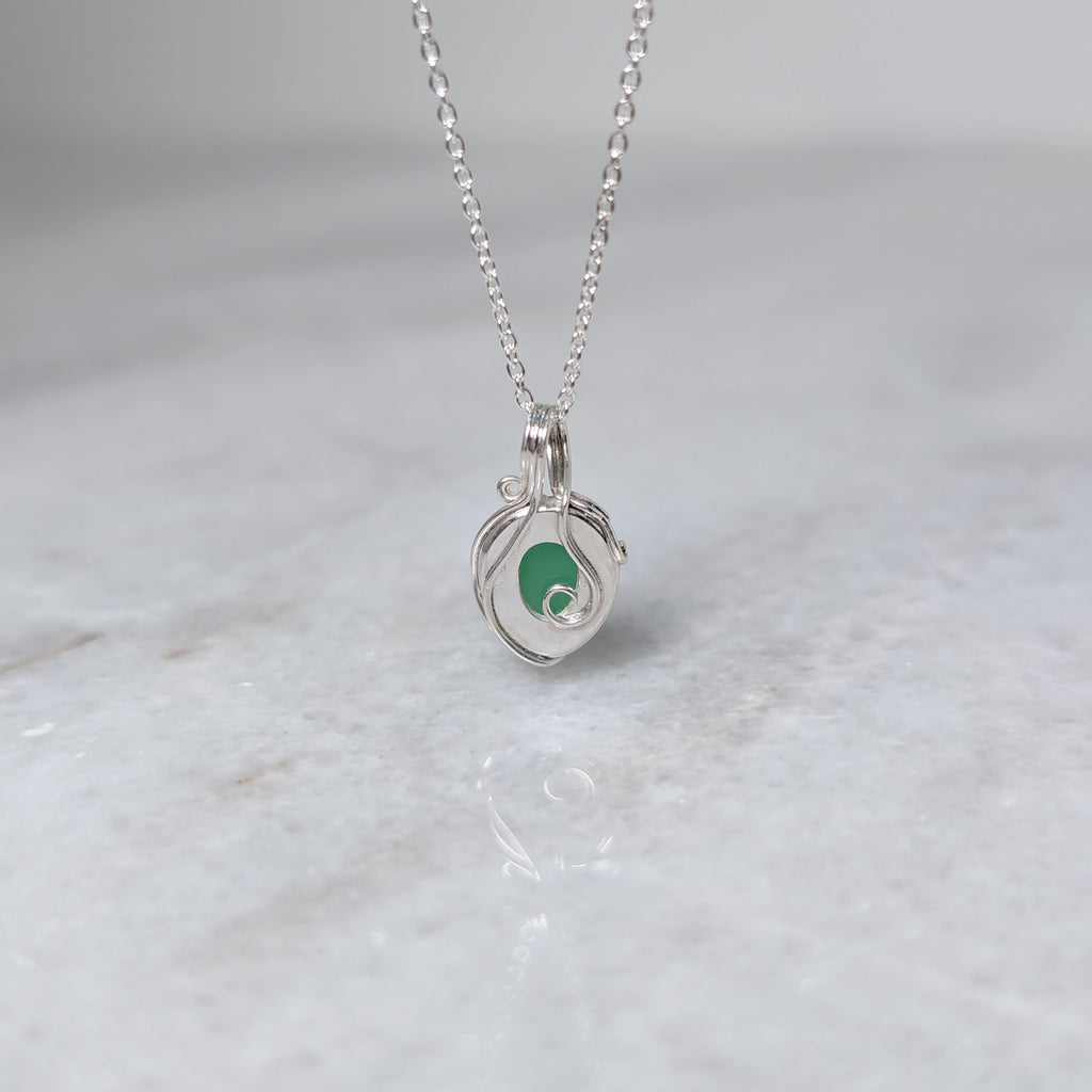 Back view of a grass green stone pendant in a silver windowed bezel with silver vines curling over it, hanging over a white marble surface