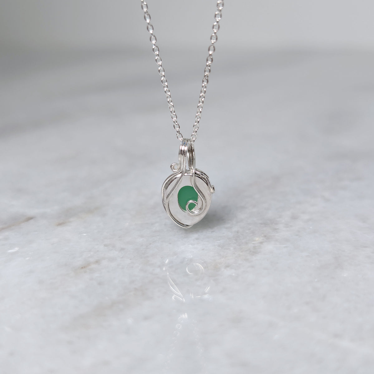 Back view of a grass green stone pendant in a silver windowed bezel with silver vines curling over it, hanging over a white marble surface