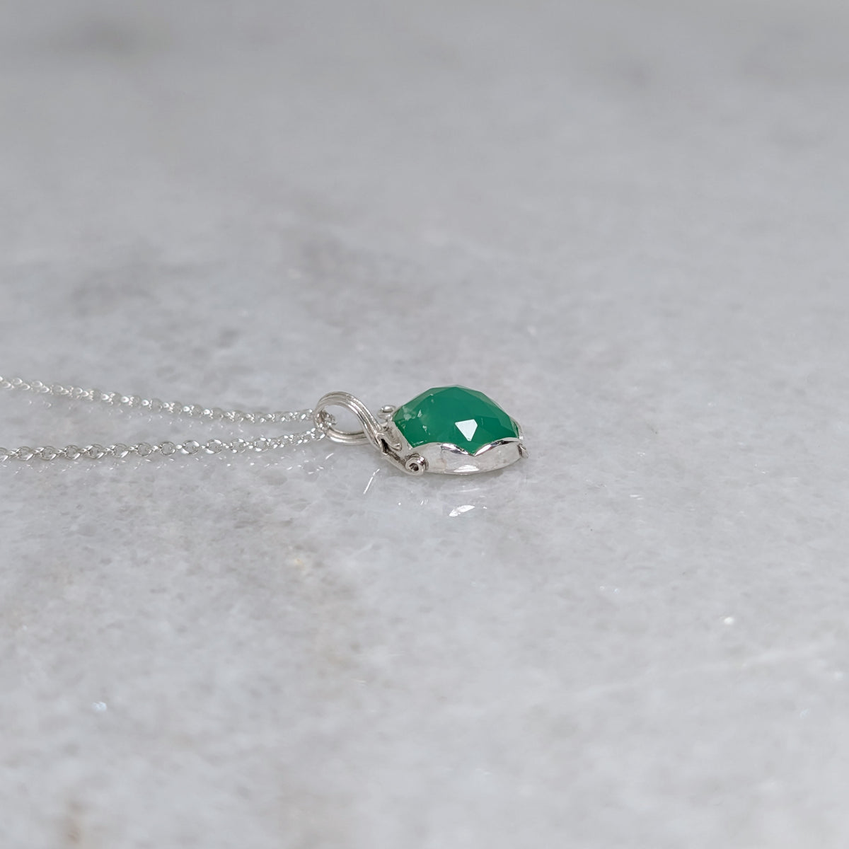 Side view of a grass green stone pendant in a silver scalloped edge bezel with curls of silver vines around the sides, on a white marble surface