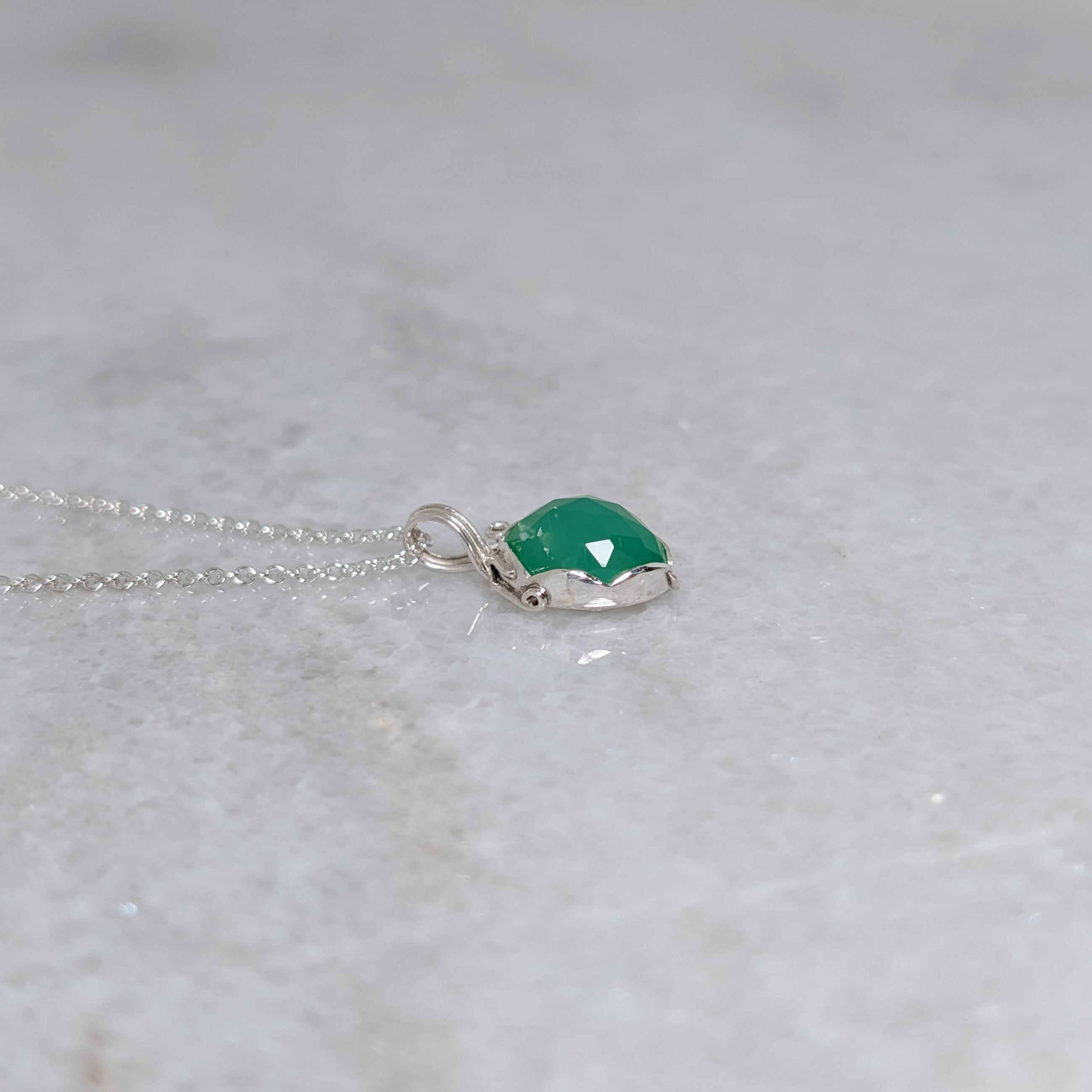 Side view of a grass green stone pendant in a silver scalloped edge bezel with curls of silver vines around the sides, on a white marble surface