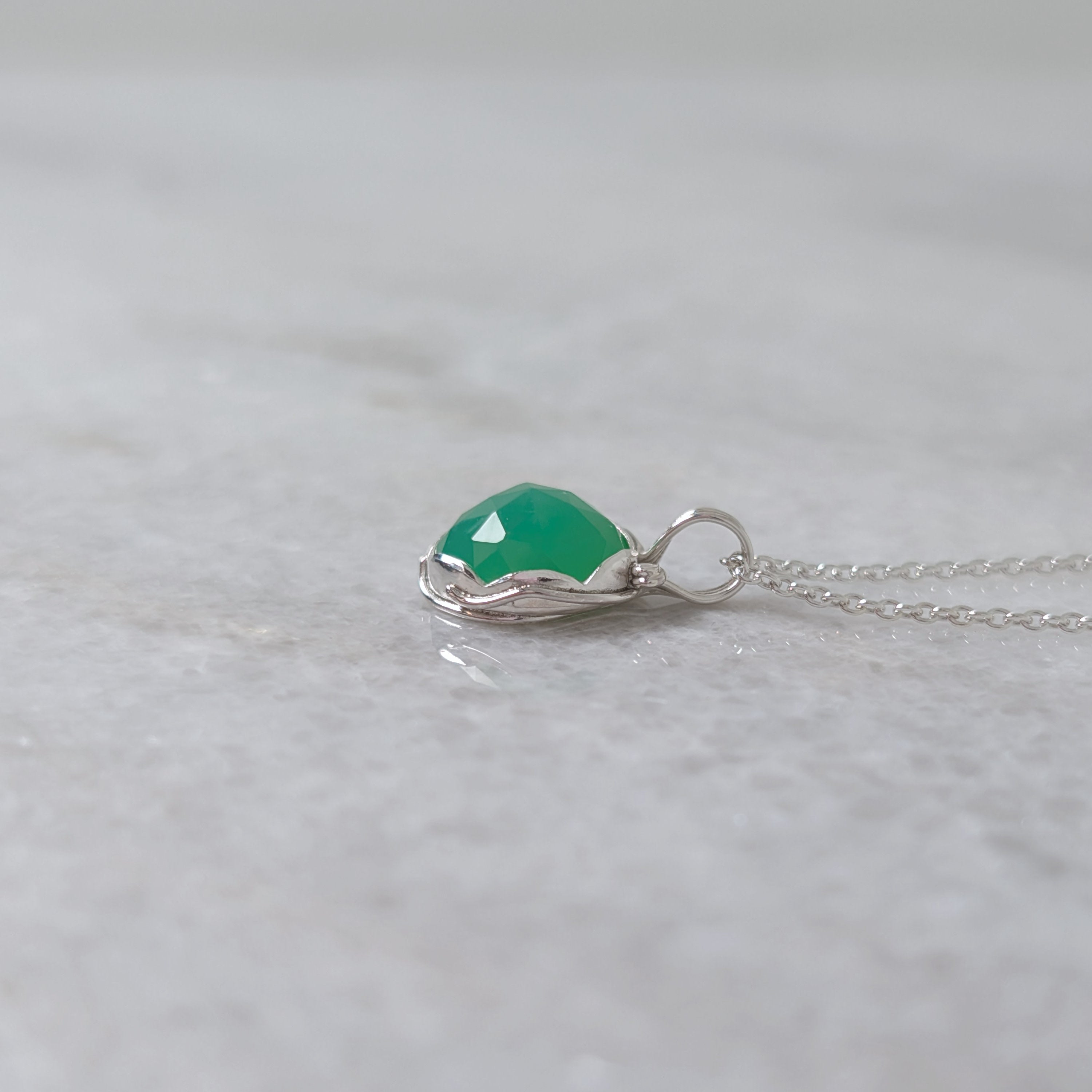 Side view of a grass green stone pendant in a silver scalloped edge bezel with silver vines around the sides, on a white marble surface
