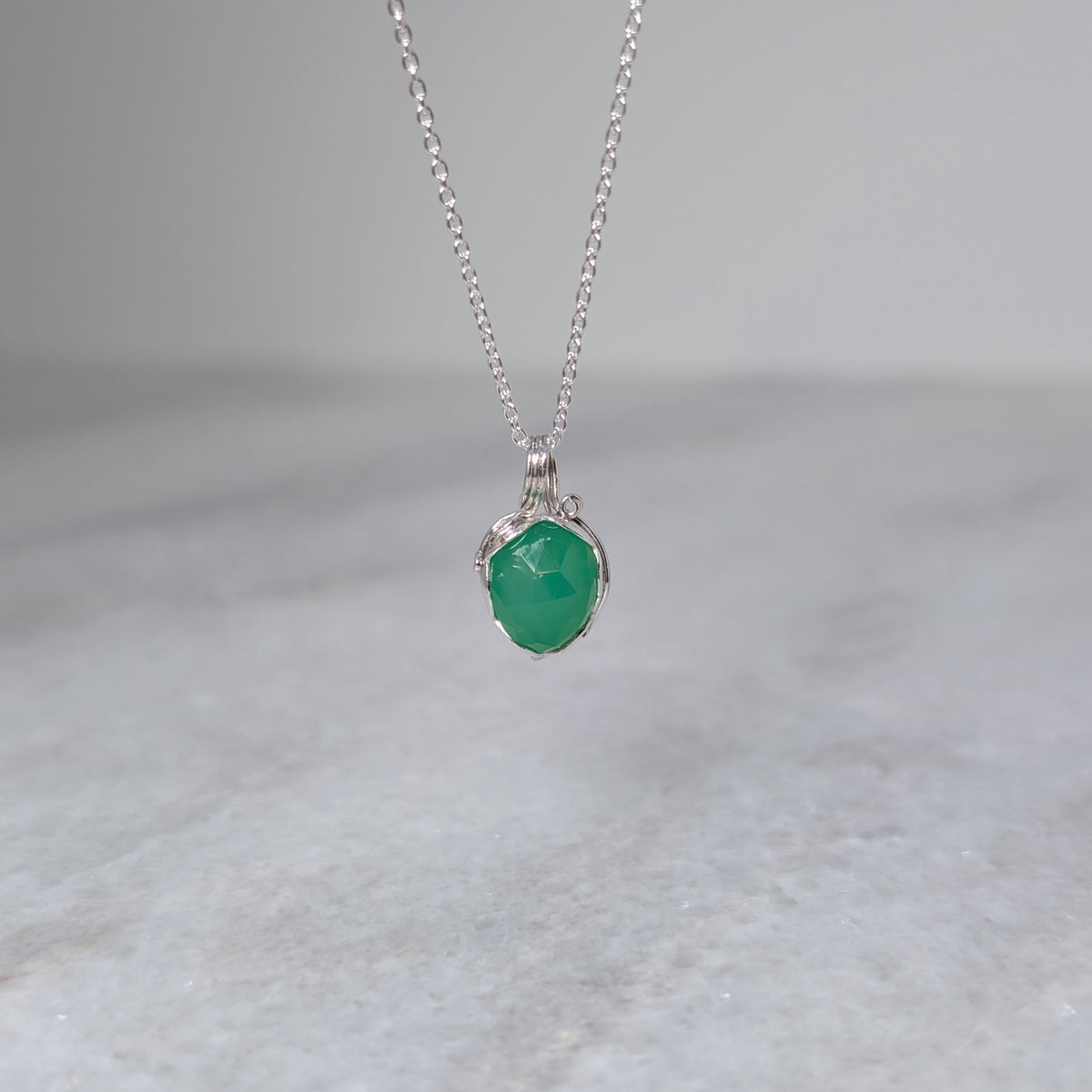 A grass green stone pendant in a silver bezel with silver vines wrapping around it, hanging over a white marble surface