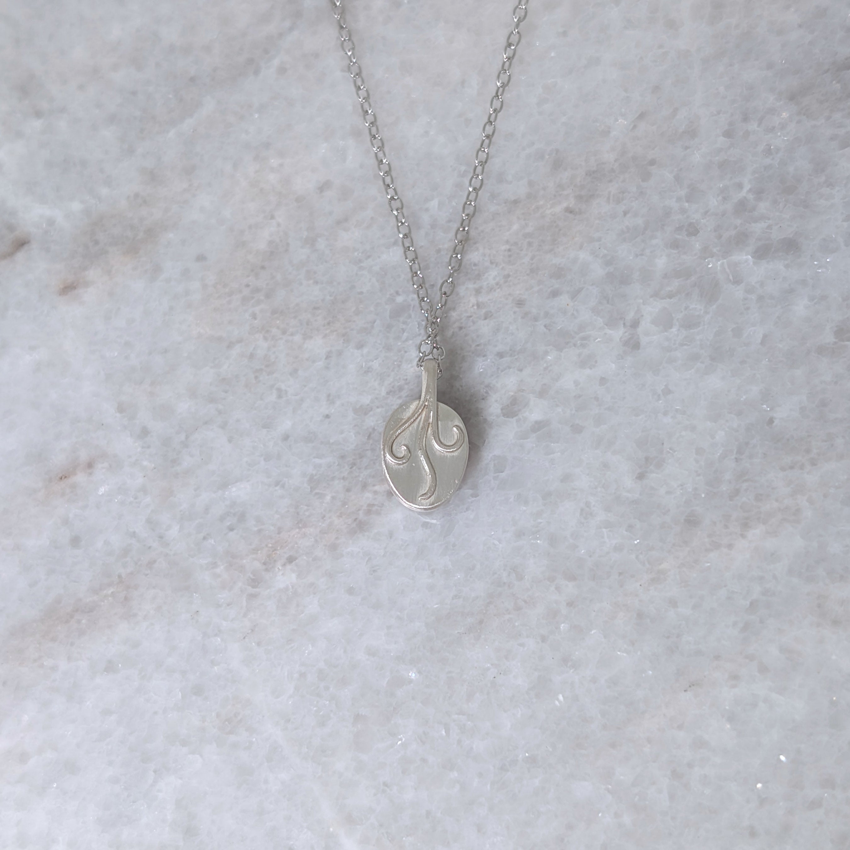 Back view of a silver necklace with three vines curling down the pendant, on a white marble surface