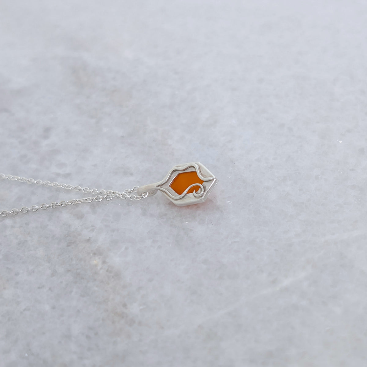 Back view of a necklace with a hexagonal orange stone set in a silver windowed bezel and wavy vine motifs framing it on a white marble background