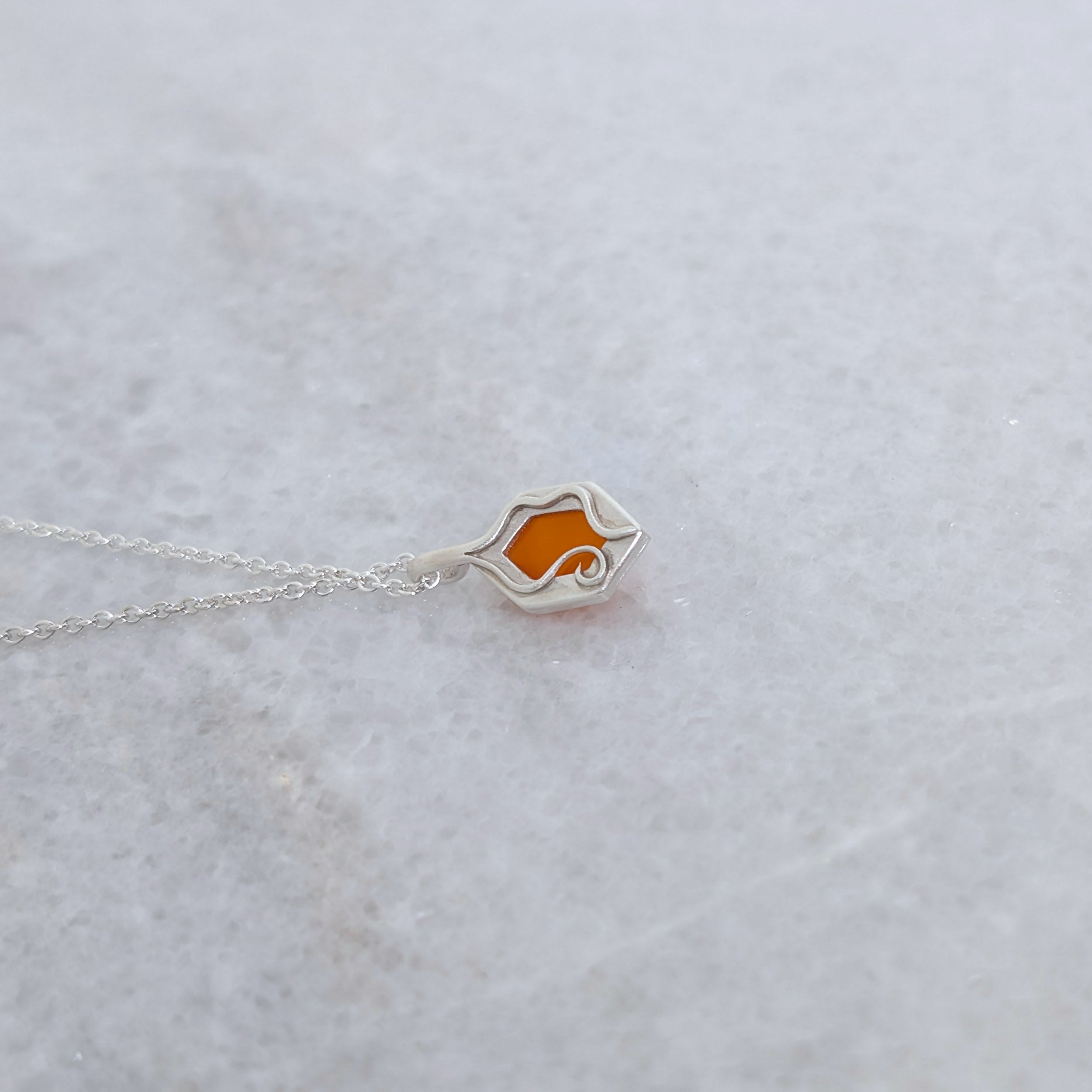 Back view of a necklace with a hexagonal orange stone set in a silver windowed bezel and wavy vine motifs framing it on a white marble background