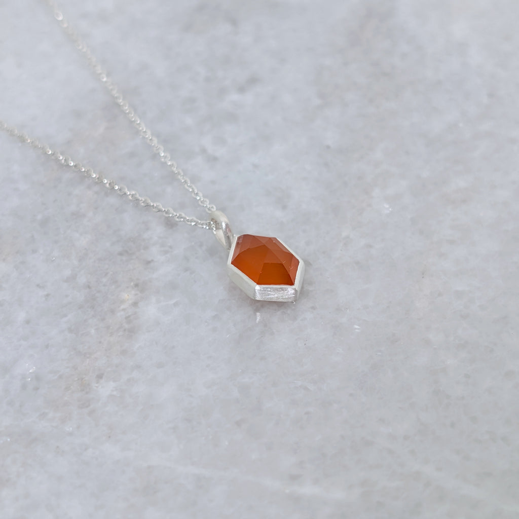 angled view of a necklace with a hexagonal orange stone set in a silver bezel on a gray background