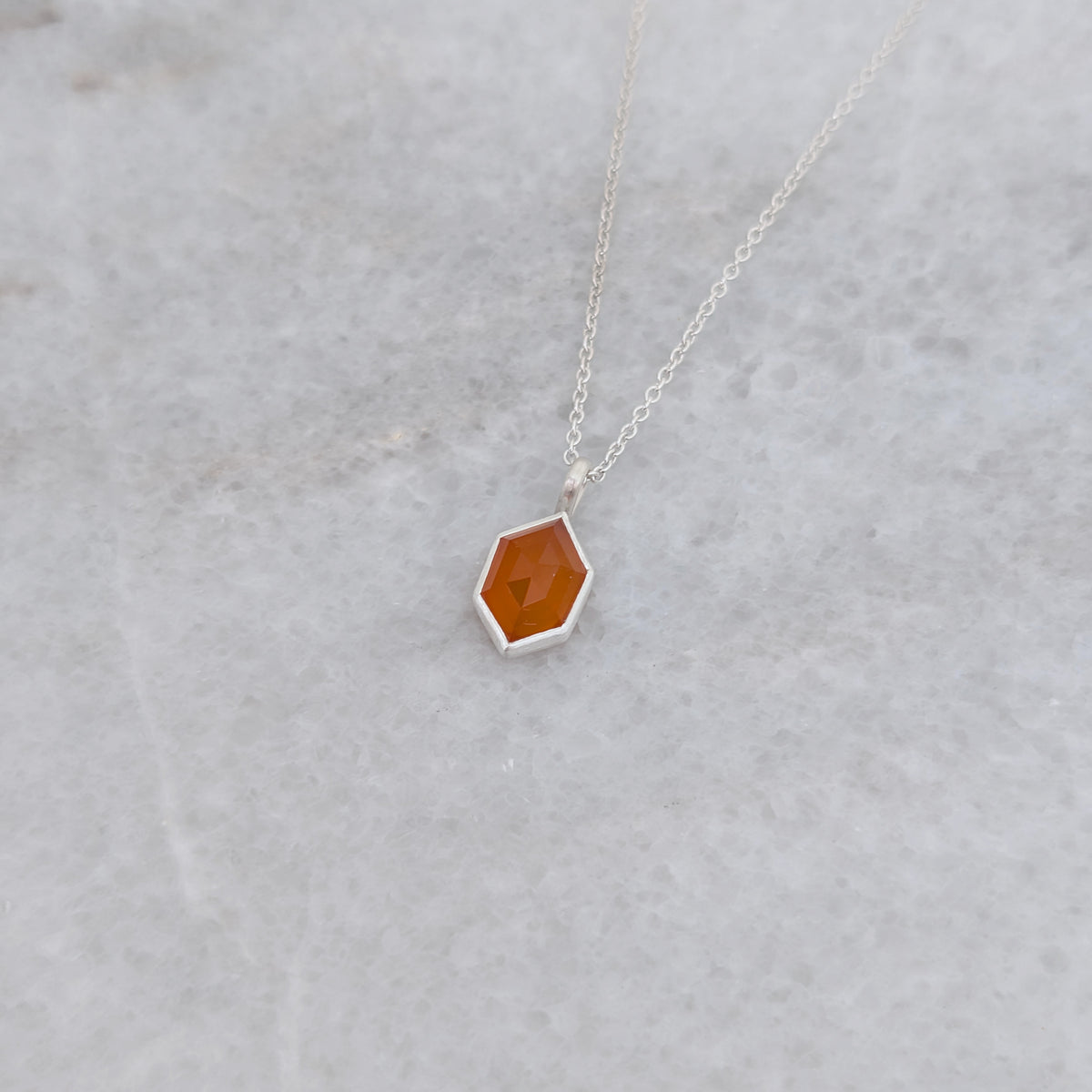 Angled view of a necklace with a hexagonal orange stone set in a silver bezel on a gray background