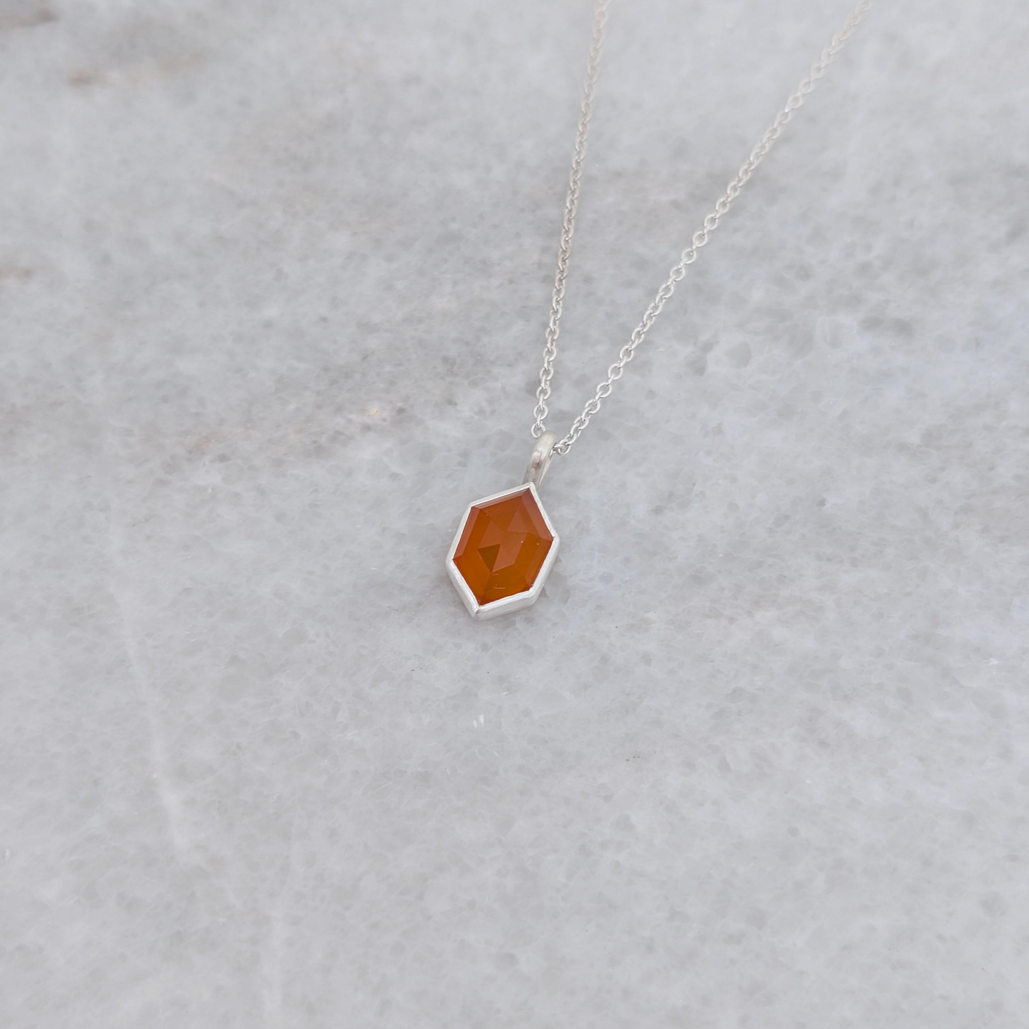 Angled view of a necklace with a hexagonal orange stone set in a silver bezel on a gray background