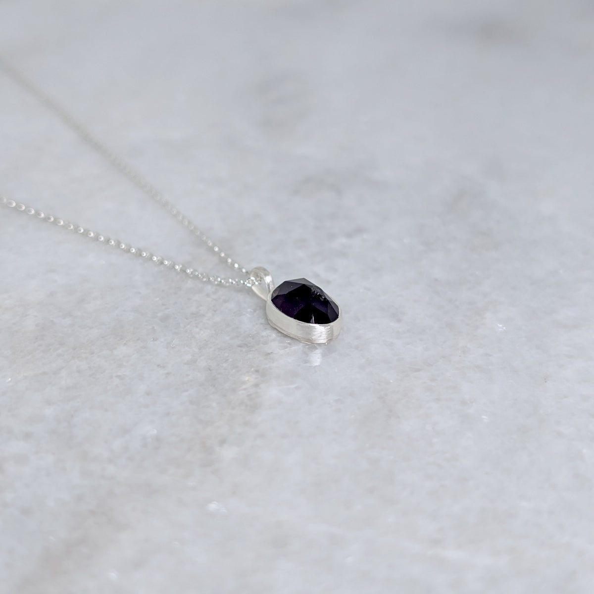 Angled view of Silver necklace with a purple stone pendant on a marble surface