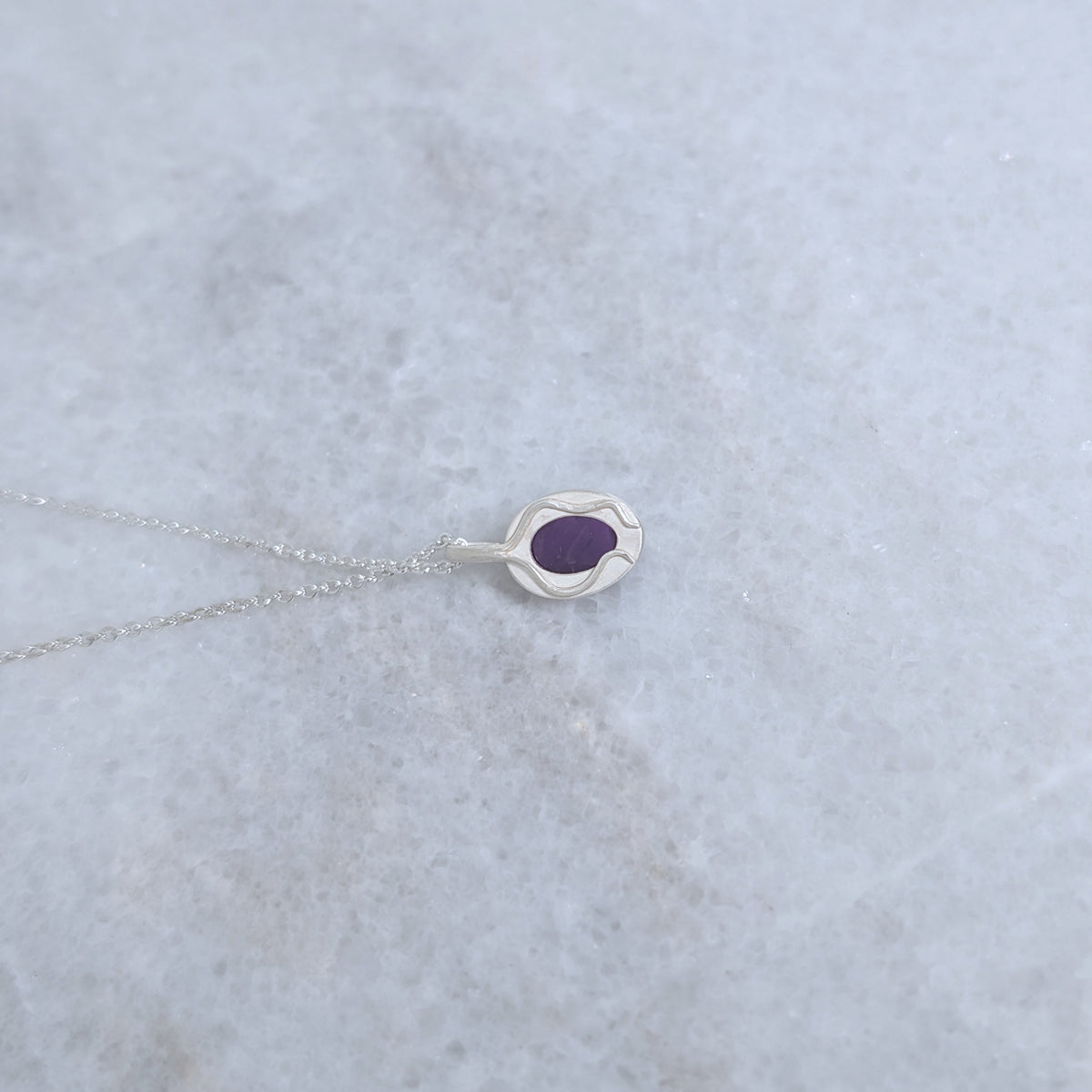 Back view of a silver necklace with a purple stone visible through a window and vine motifs framing it, on a marble surface