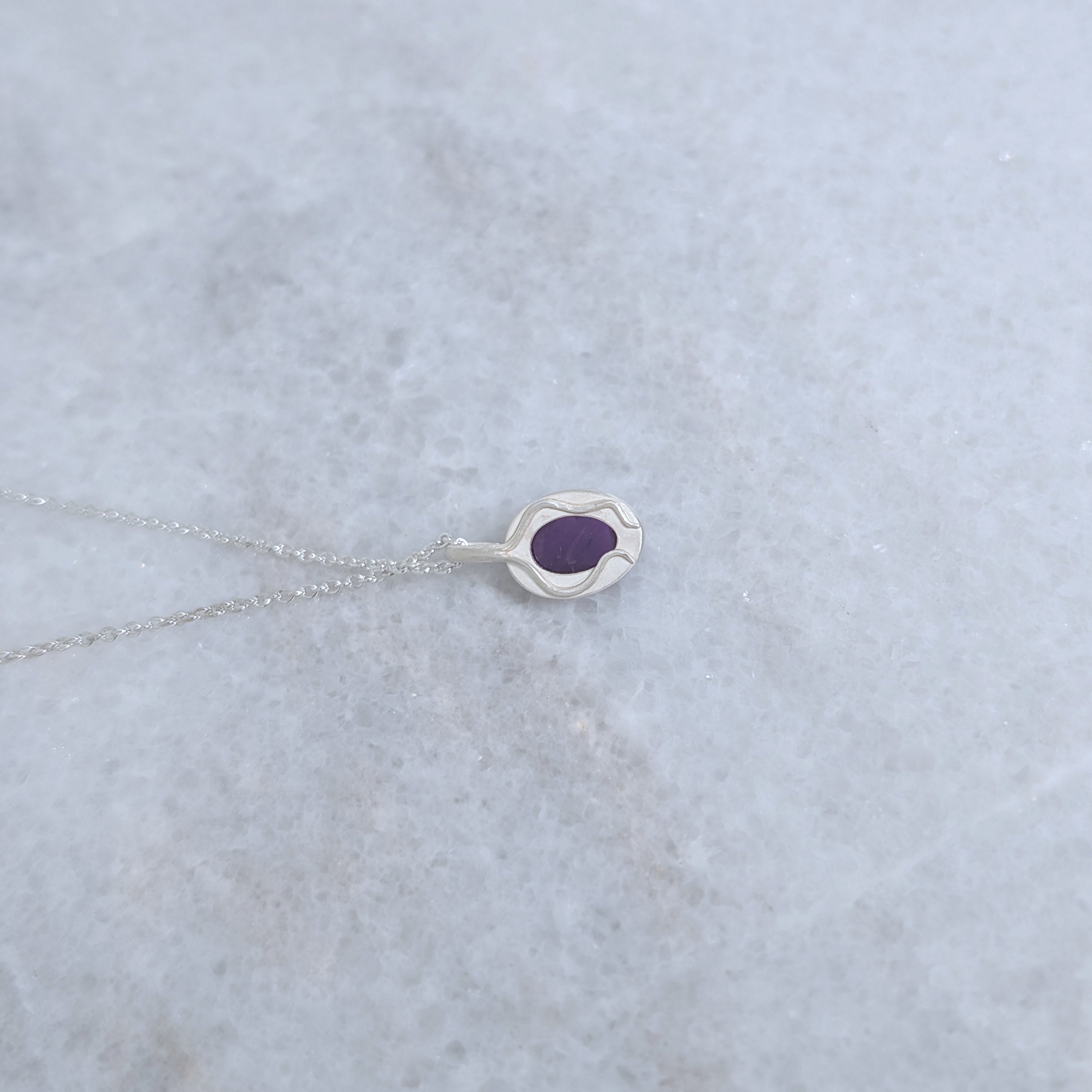 Back view of a silver necklace with a purple stone visible through a window and vine motifs framing it, on a marble surface