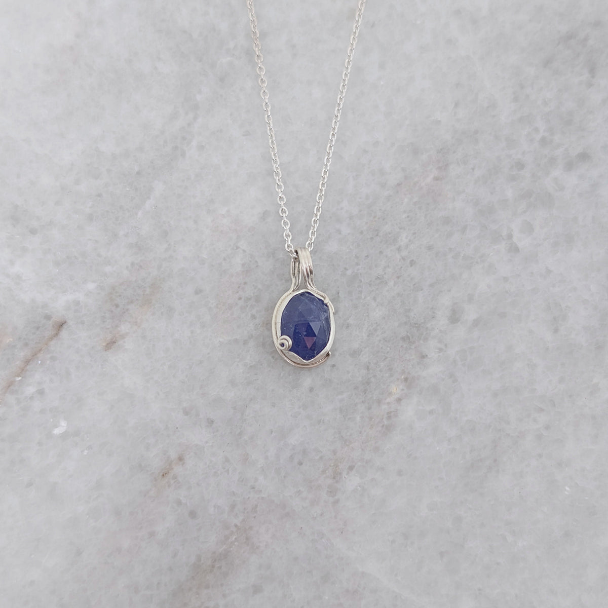 Silver necklace with a blueish purple pendant with vine motifs on a white marble surface