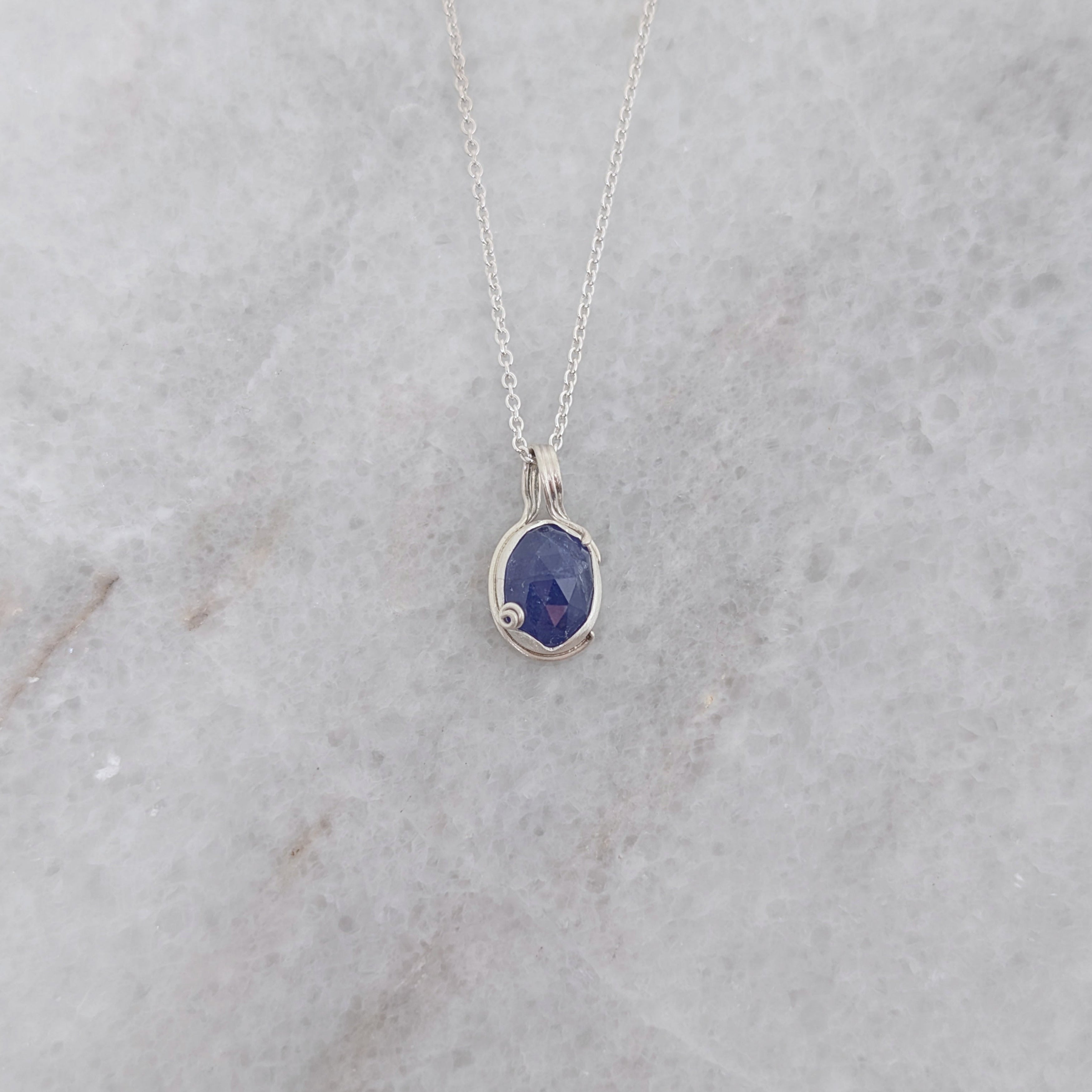 Silver necklace with a blueish purple pendant with vine motifs on a white marble surface