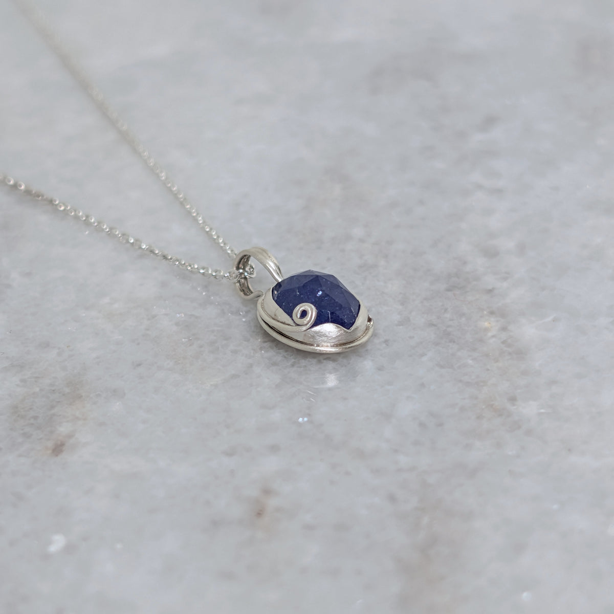 Angled view of Silver necklace with a blueish purple pendant with curled vine motif on a white marble surface