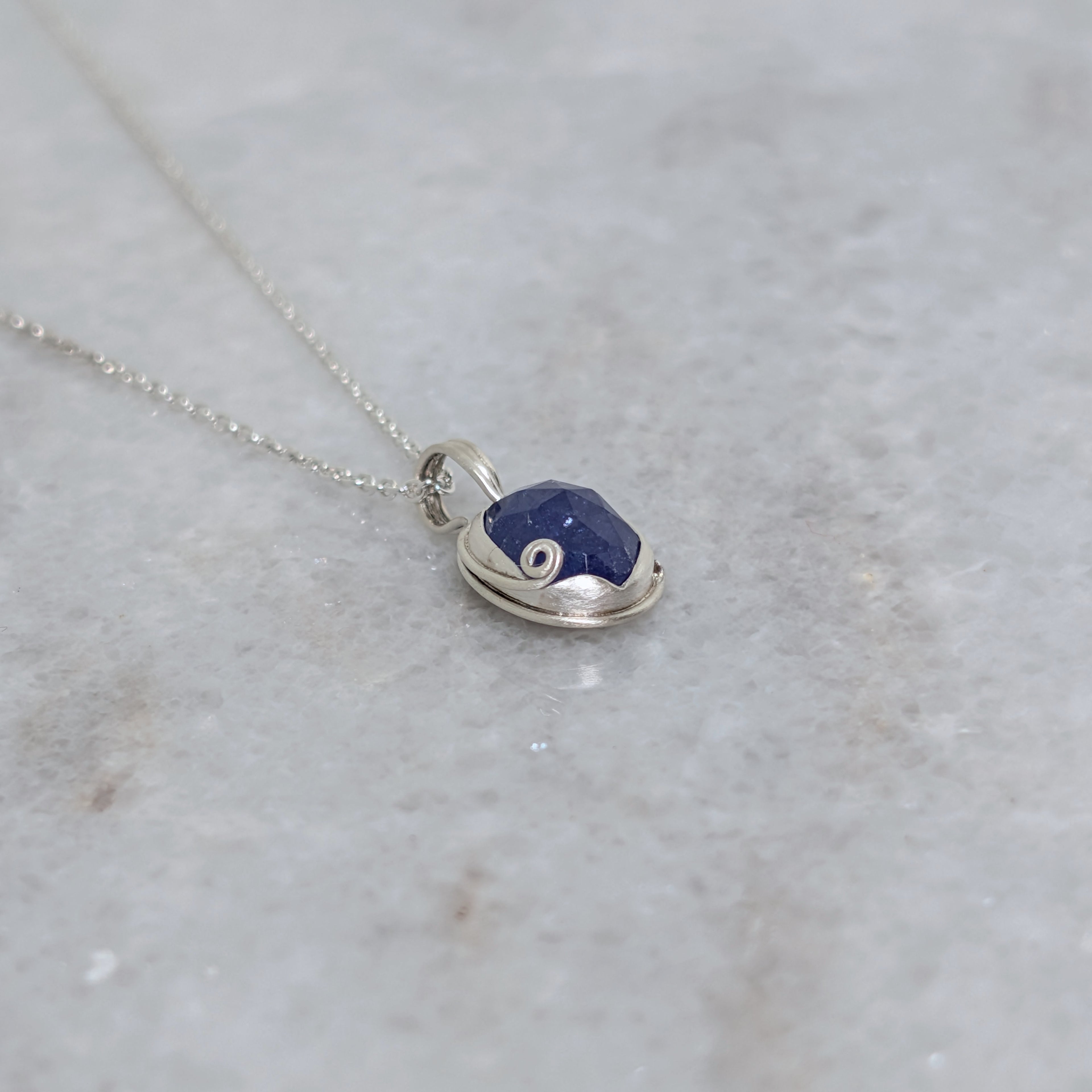Angled view of Silver necklace with a blueish purple pendant with curled vine motif on a white marble surface