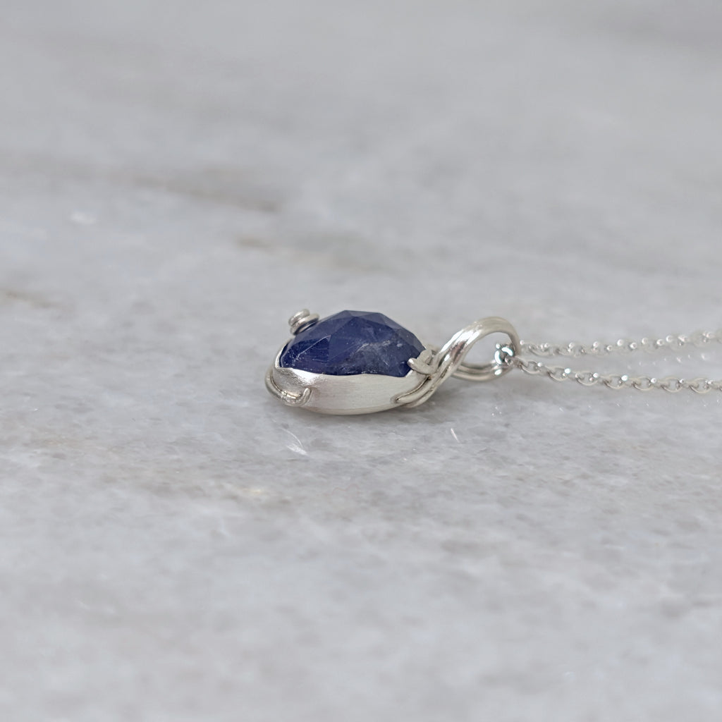 Side view of a silver pendant with a bluish purple stone and vine motifs on a white marble surface