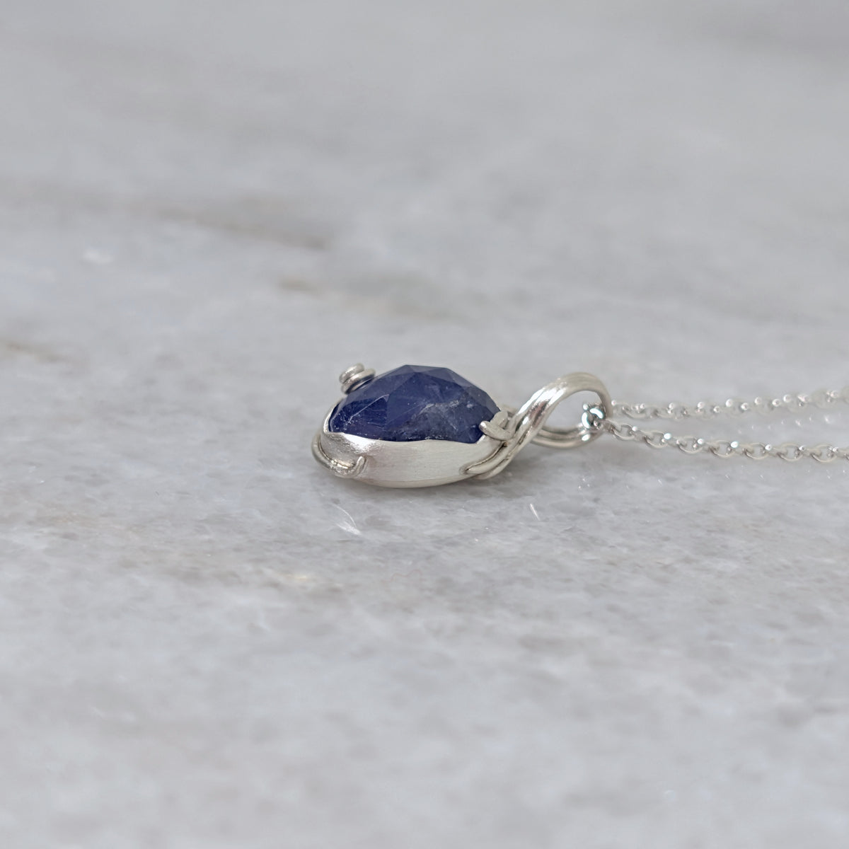 Side view of a silver pendant with a bluish purple stone and vine motifs on a white marble surface