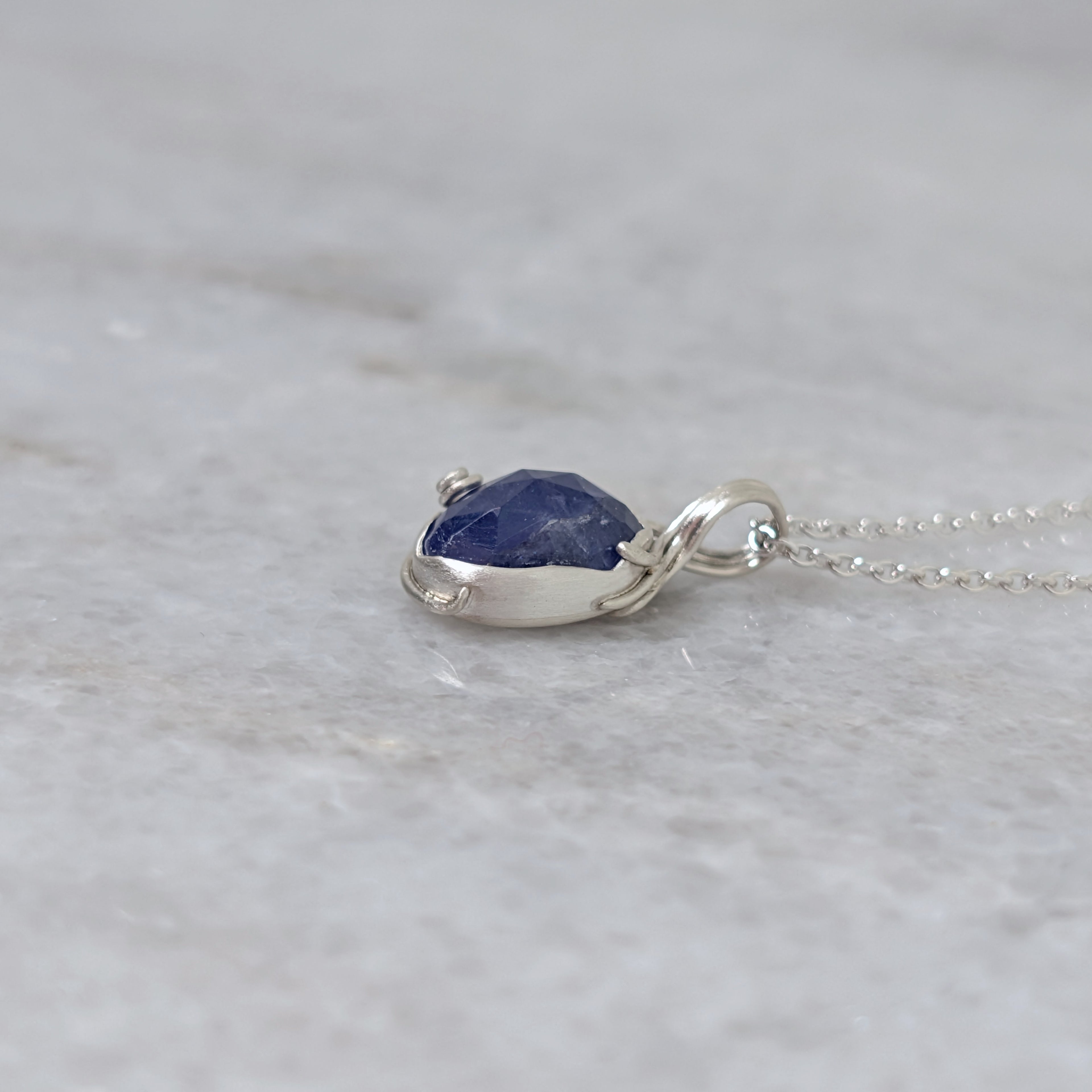 Side view of a silver pendant with a bluish purple stone and vine motifs on a white marble surface
