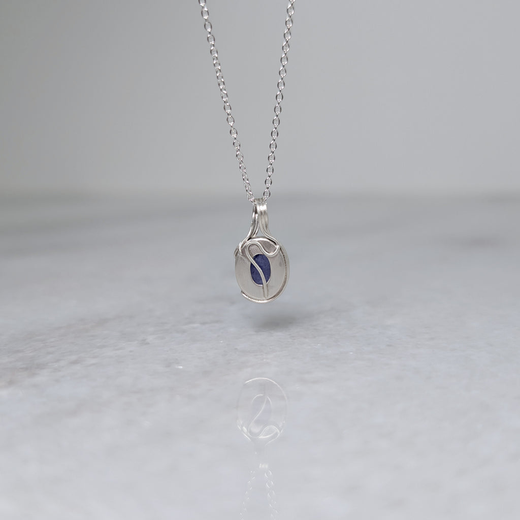 Back view of a silver necklace with a bluish purple stone and a wavy vine motif across the back, hanging in front of a white background
