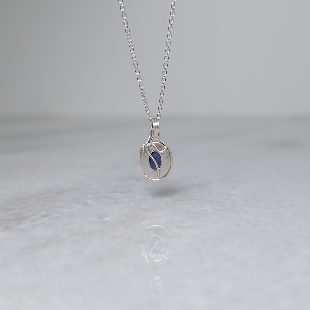 Back view of a silver necklace with a bluish purple stone and a wavy vine motif across the back, hanging in front of a white background