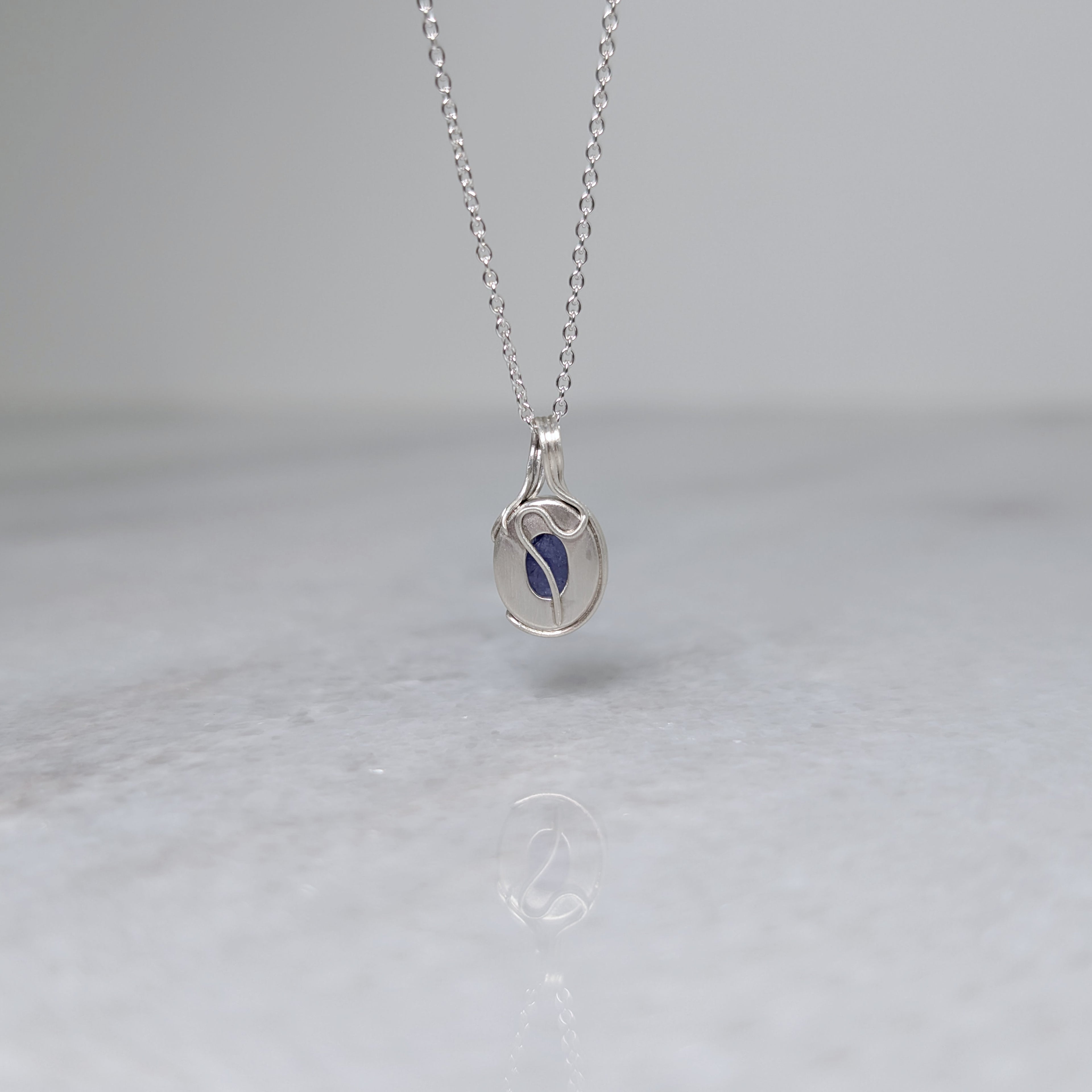 Back view of a silver necklace with a bluish purple stone and a wavy vine motif across the back, hanging in front of a white background