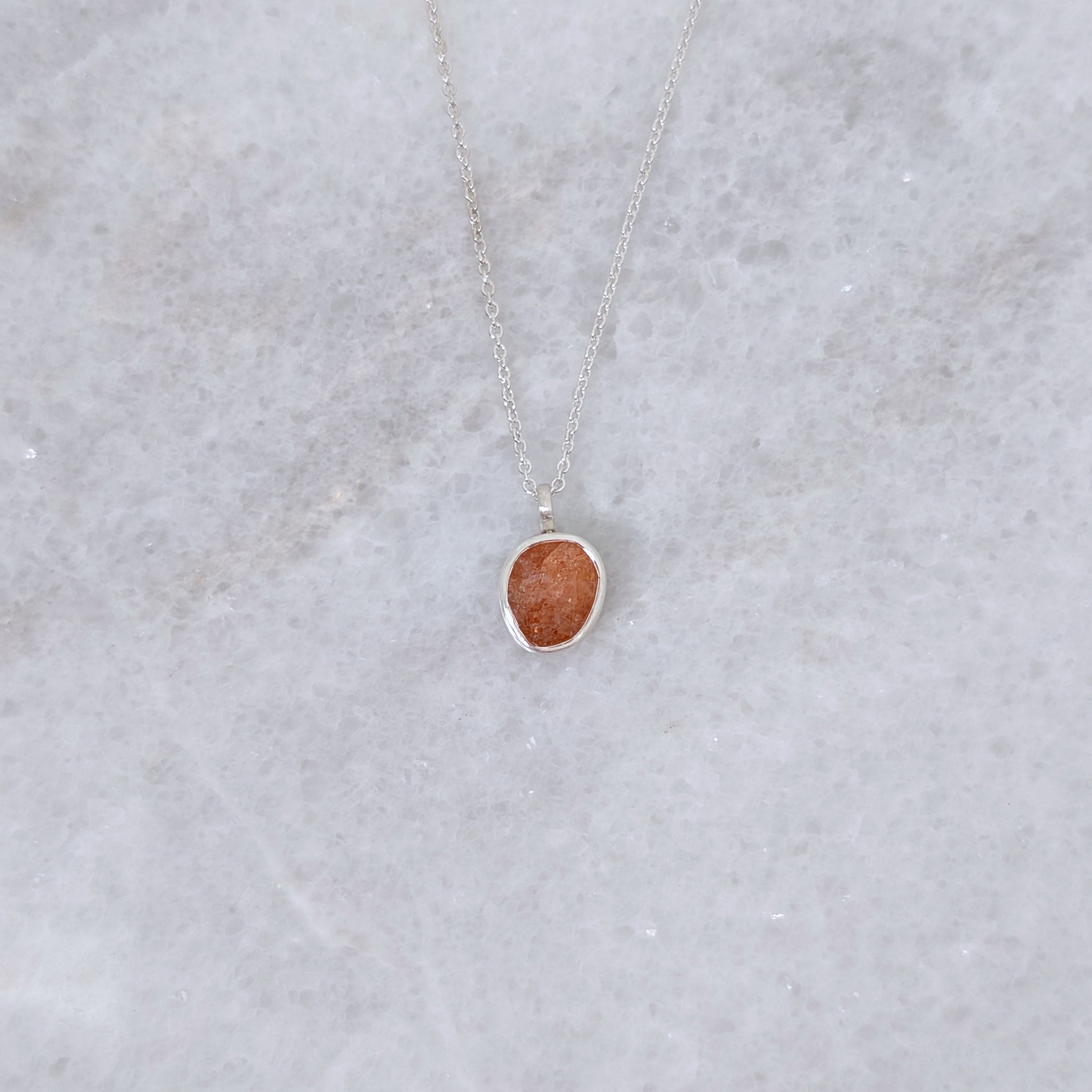 Necklace with a speckled orange stone set in a silver bezel on a white marble backround