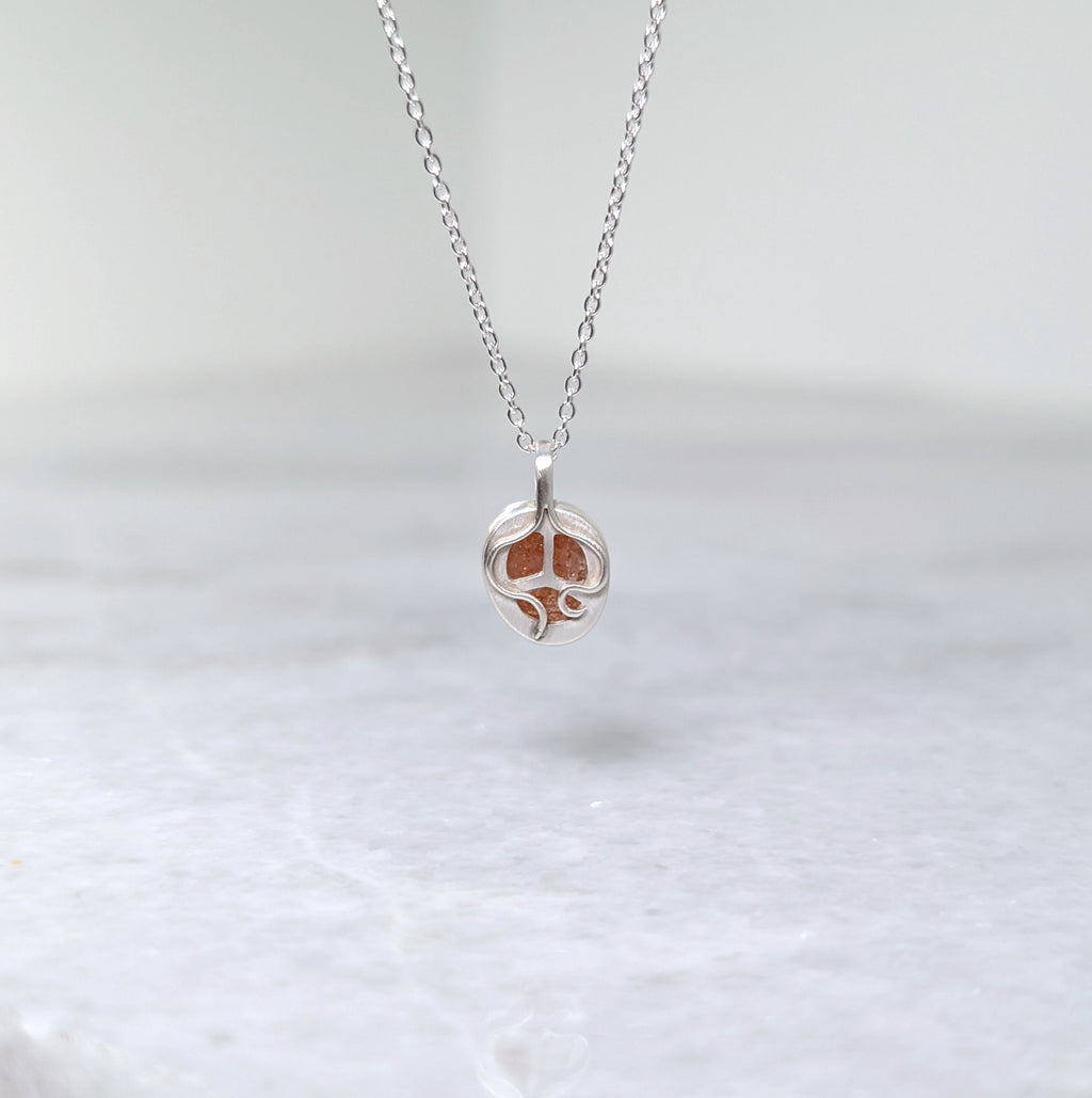 The back side of a silver pendant with an orange speckled stone visible through windows framed by silver vine motifs