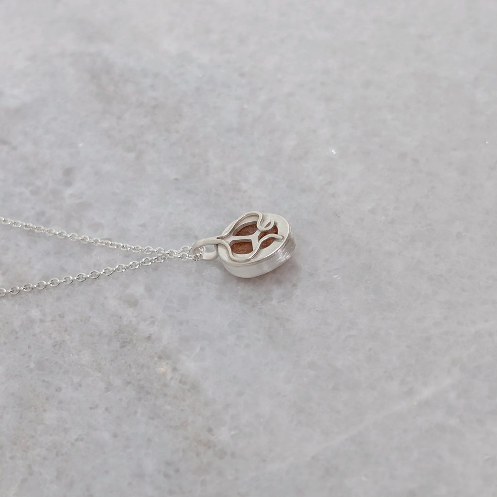 The back side of a silver necklace with an orange speckled stone visible through windows framed by silver vine motifs