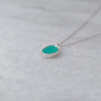 Angled view of a silver necklace with a teal colored stone in a silver bezel on a white marble background