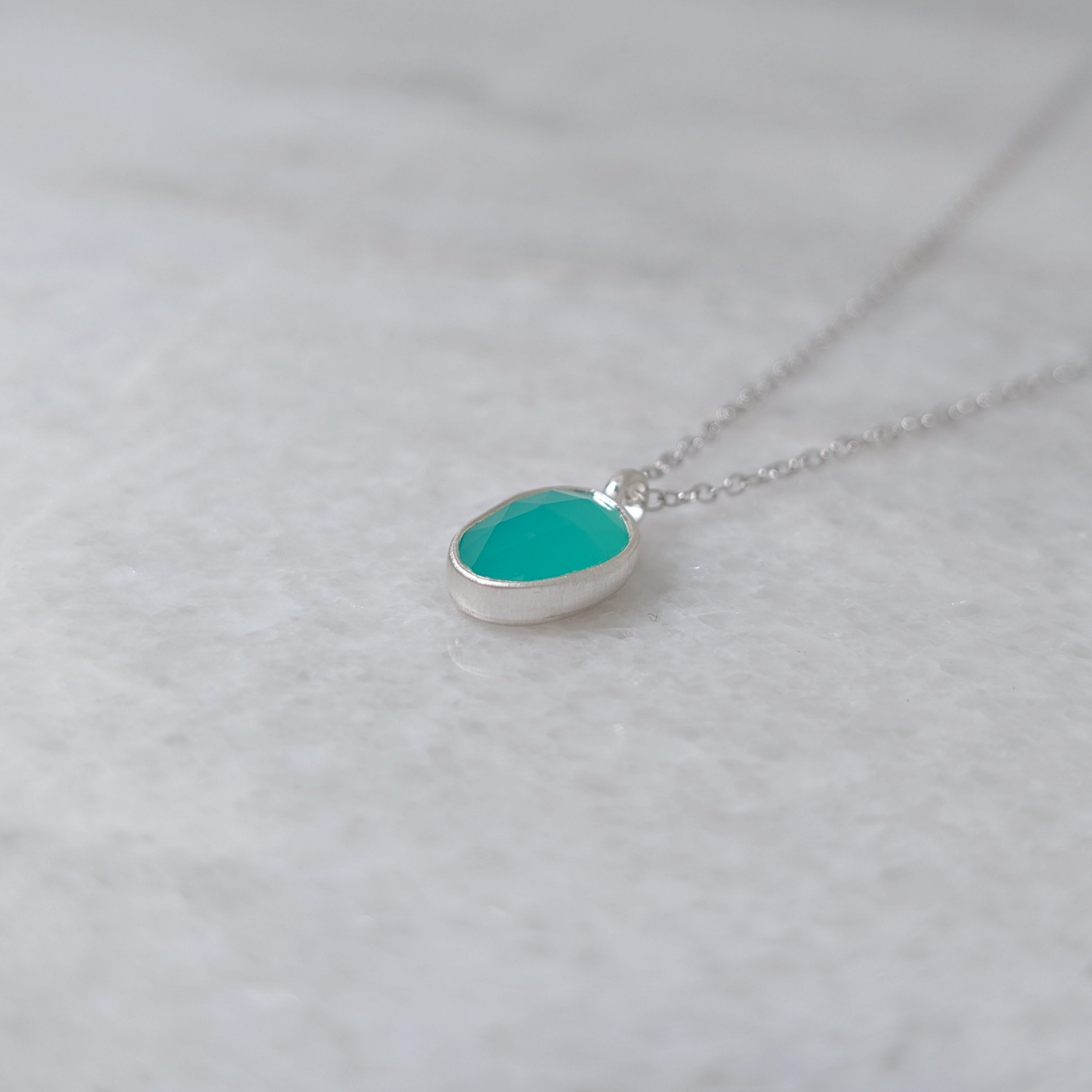 Angled view of a silver necklace with a teal colored stone in a silver bezel on a white marble background