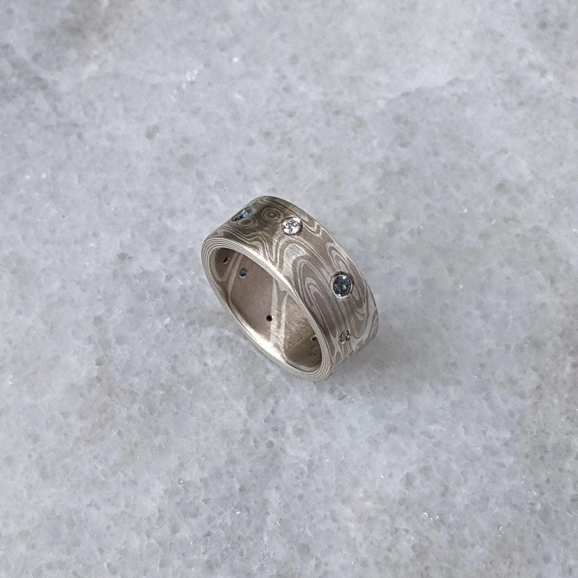 A mokume gane ring made of silver and white gold with a wavy wood grain pattern, with scattered stones set flush.