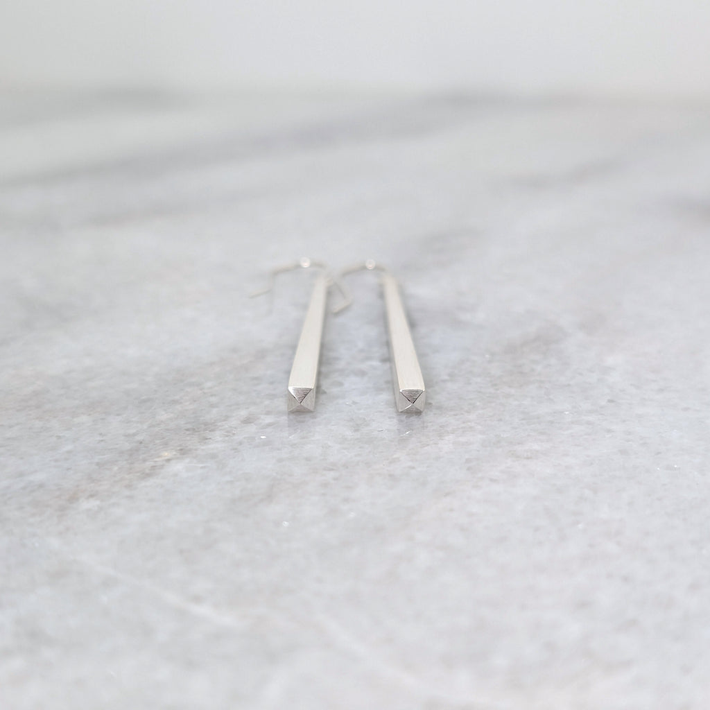 Faceted end detail of Silver tapered bar earrings on a white marble surface